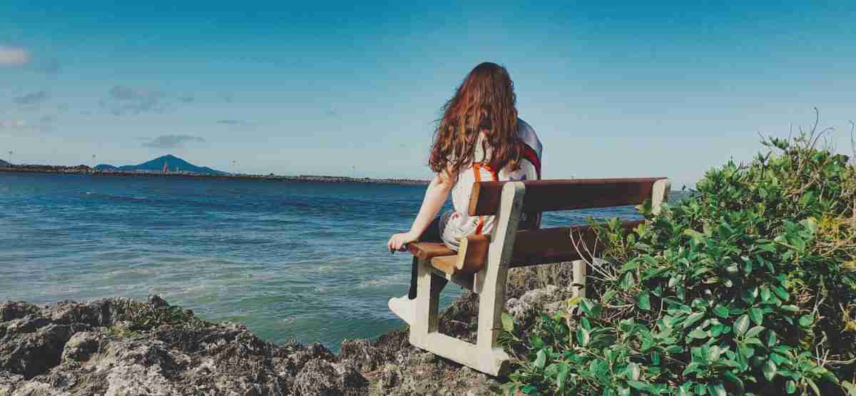 woman wearing white jacket sitting on white wooden bench facing body of water viewing mountain under blue and white sky