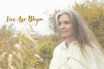 Live Age Bloom Begins Here- A woman with long hair and partly grey in a field.